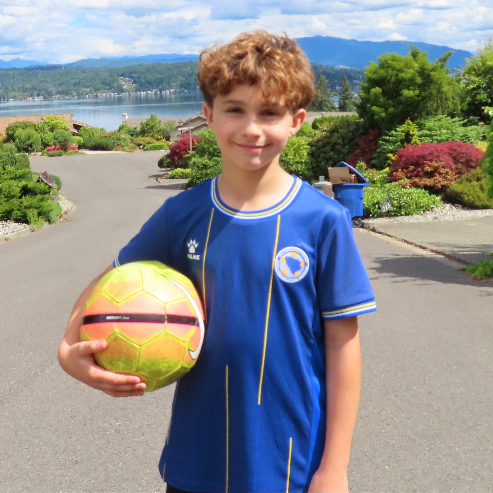 Child holding a colorful ball on a residential street with greenery and mountains in the background